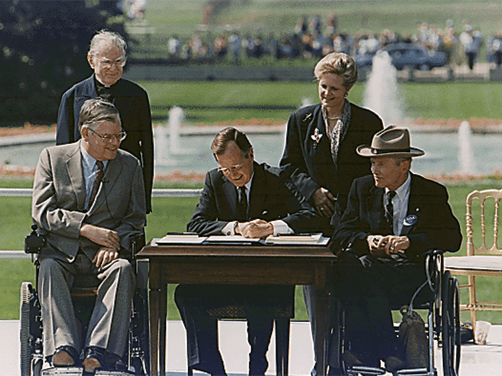 President George H.W. Bush sitting outdoors at a table signing the Americans with Disabilities Act. His wife and three men surround him. Two of the men are in wheelchairs.